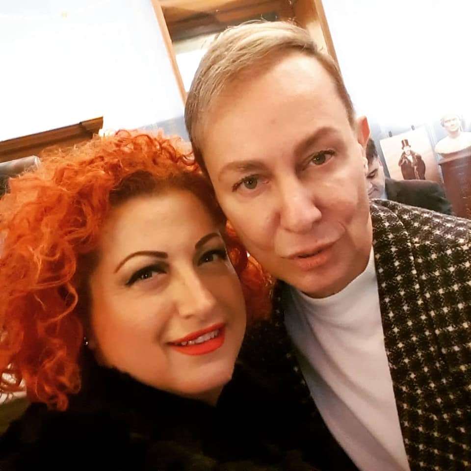 Δύο άτομα ποζάρουν κοντά το ένα στο άλλο, με έντονα χαρακτηριστικά και χαμογελαστά πρόσωπα, σε ένα κοινωνικό ή δημόσιο περιβάλλον.