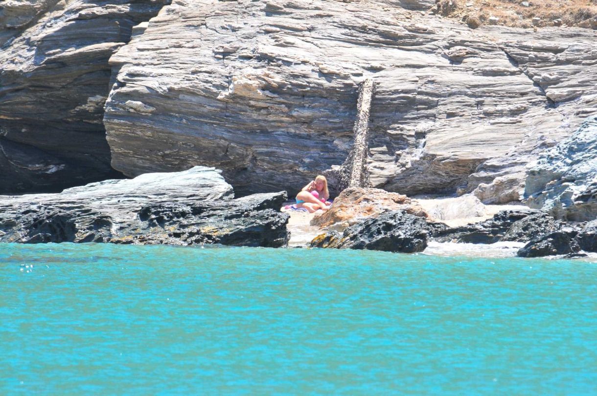 A woman in a swimsuit sitting on rocks by the sea, with cliffs in the background, enjoying a sunny day.