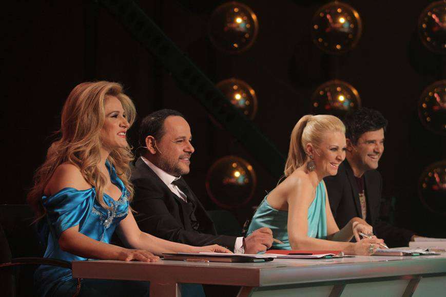 Τέσσερις κριτές σε τηλεοπτικό show, καθισμένοι στο τραπέζι αξιολόγησης, με φωτισμένα φόντα.