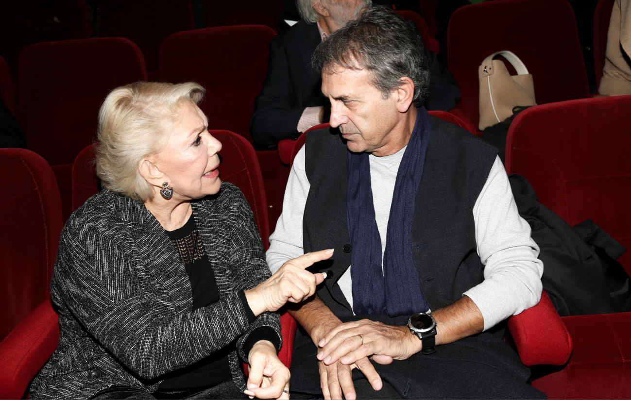 Δύο άτομα συζητούν σε θέατρο, με θηλυκό πρόσωπο να δείχνει προς τον άνδρα, που φορά ρούχα σε σκούρες αποχρώσεις.