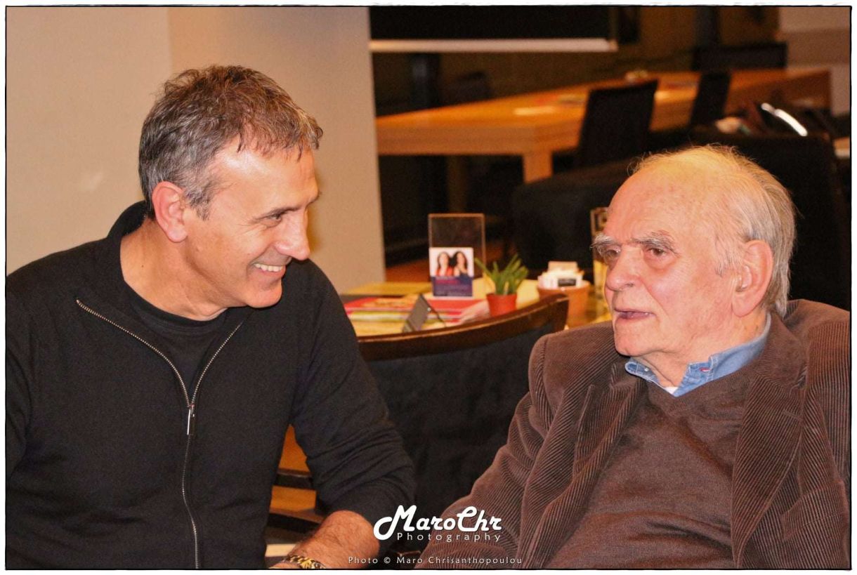 Δύο άνδρες καθισμένοι σε έναν καφέ, συζητούν σε φιλική ατμόσφαιρα, με χαμόγελα και εκφράσεις ευχαρίστησης.