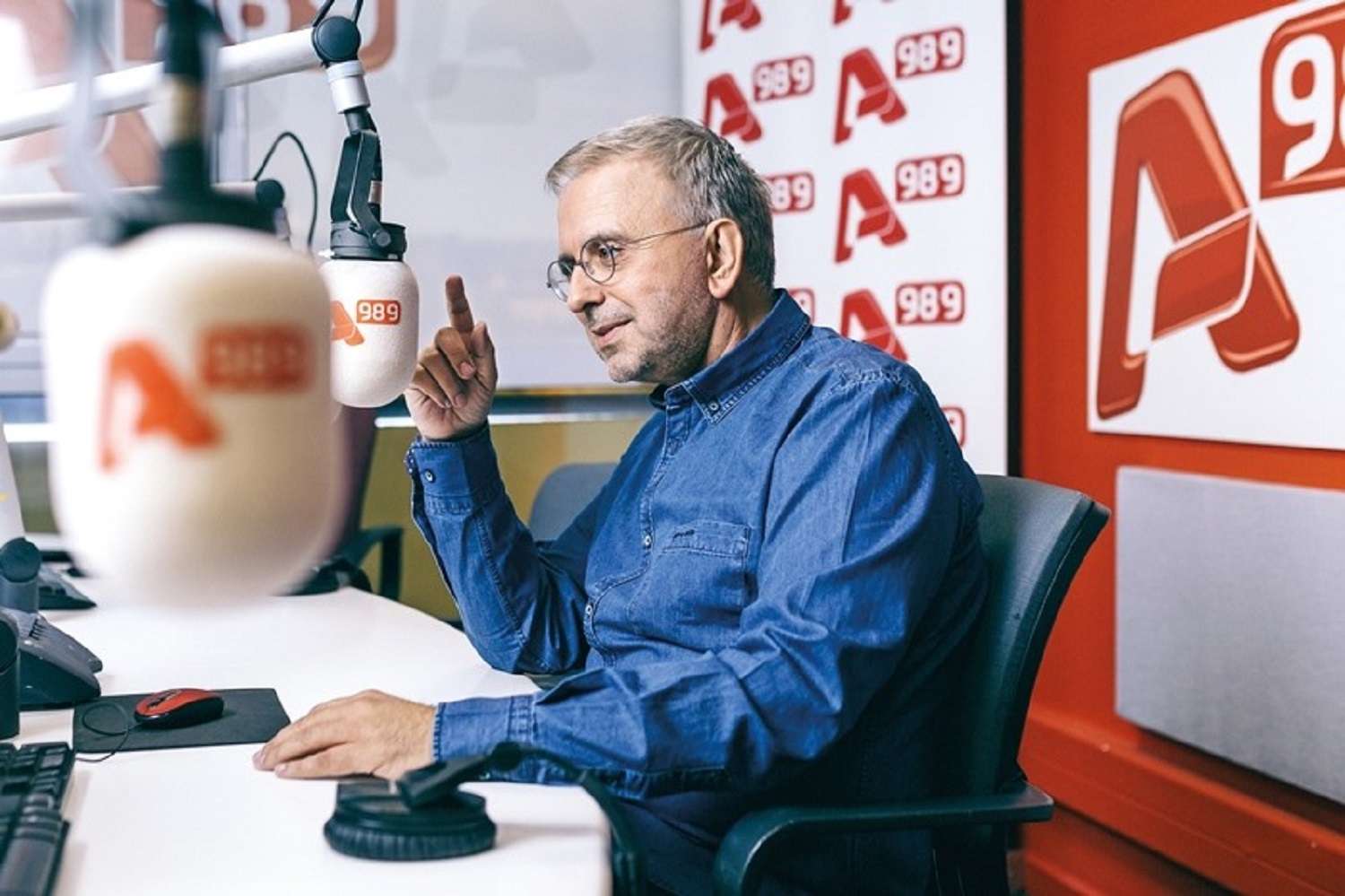 Ο Δήμος Βερύκιος στο στούντιο ραδιοφωνικού σταθμού, φοράει πουκάμισο και μιλάει στο μικρόφωνο.