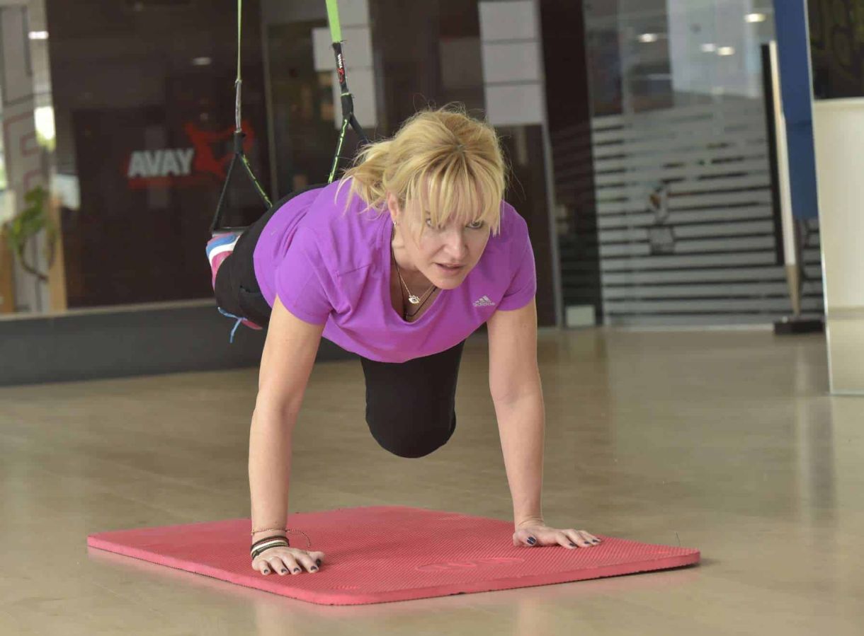 Γυναίκα που εκτελεί push-ups με χρήση ιμάντων σε γυμναστήριο, με έντονη έκφραση προσώπου.