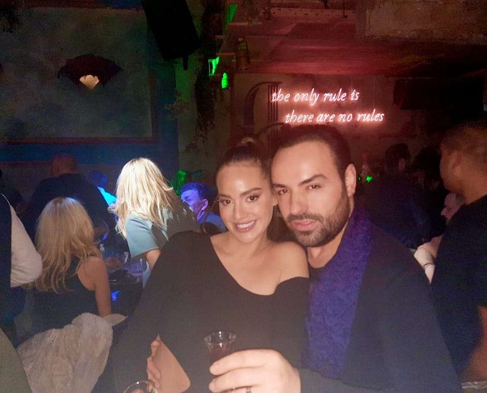 Η Ρία Ελληνίδου ποζάρει χαμογελαστή με έναν άνδρα σε μια νυχτερινή έξοδο, κρατώντας ποτήρια ενώ πίσω τους διακρίνεται κόσμος και φωτισμός από κλαμπ.