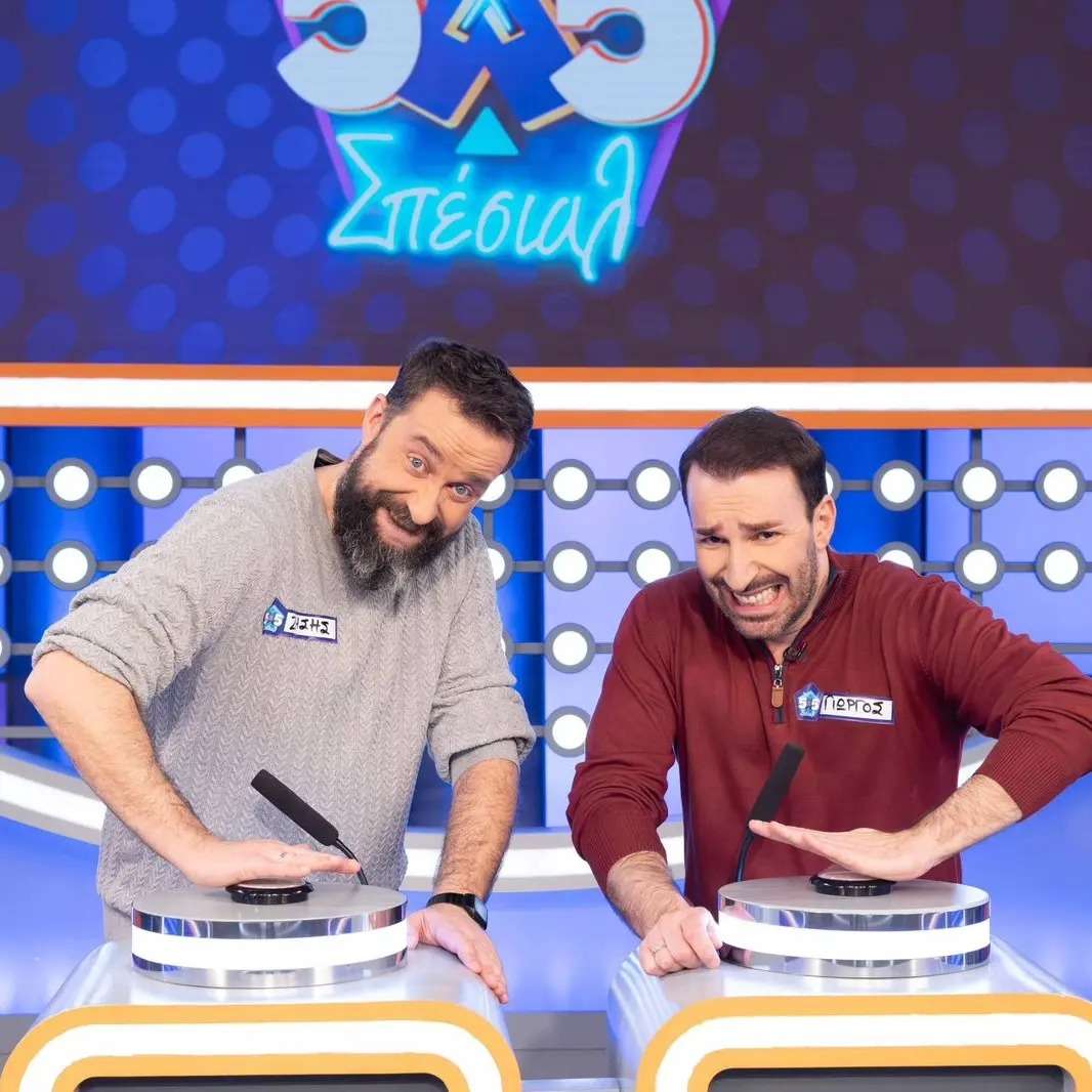 Δύο άνδρες συμμετέχουν σε τηλεοπτικό παιχνίδι, στέκονται μπροστά από στιγμιαίους υποδοχείς και φαίνεται να περιμένουν να απαντήσουν. Ο φωτισμός της σκηνής είναι ζωντανός και το λογότυπο της εκπομπής είναι ορατό στο φόντο.